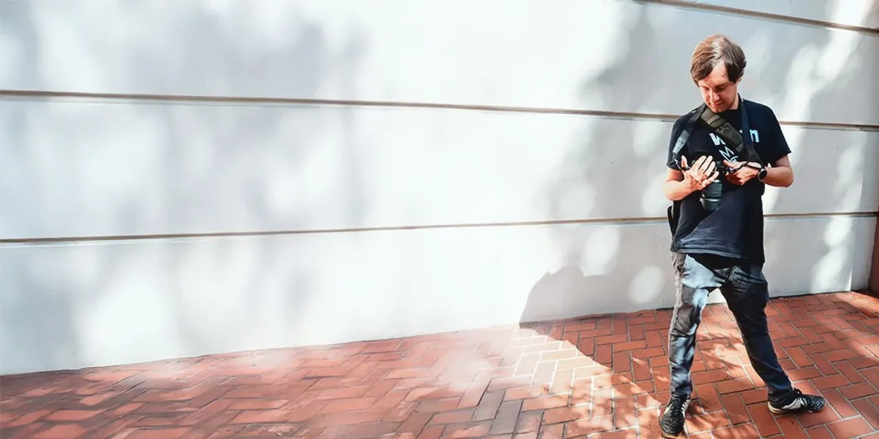 A man standing on a red brick floor, surrounded by white walls, with shadows cast on the wall behind him.